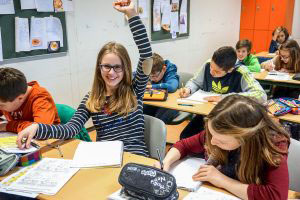 Blick ins Klassenzimmer. Mehrere Schüler schreiben in ihre Hefte. Eine Schülerin streckt ihren Arm und blickt lächelnd in die Kamera.