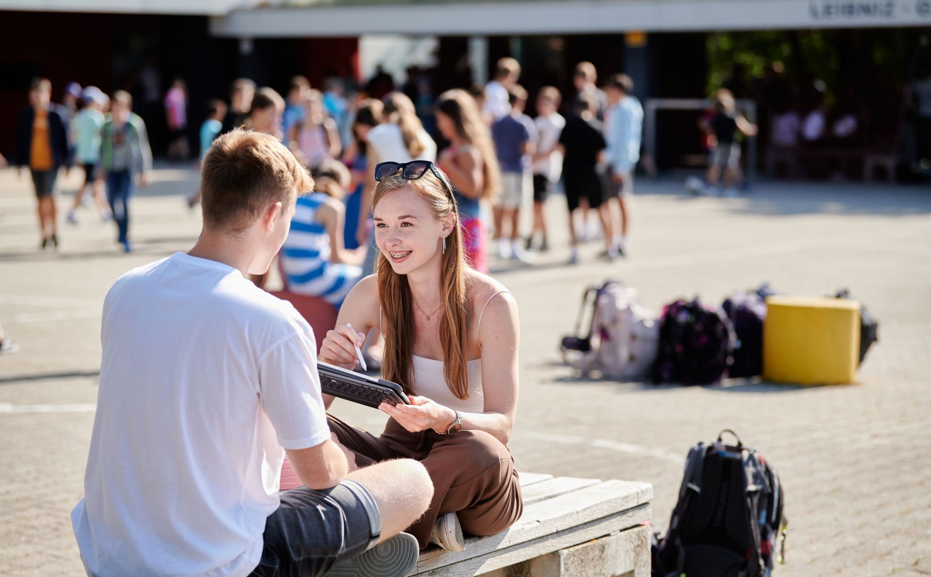 Eine Schülerin und ein Schüler sitzen auf dem Pausenhof auf einer Bank und haben ein Tablet zwischen sich.